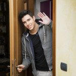 35114849 - smiling young man getting out of door waving at the camera with a friendly cheerful smile as he peers around the edge of a wooden door