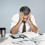 Man at desk in shirt and tie holding his head and worrying about money and the economy.